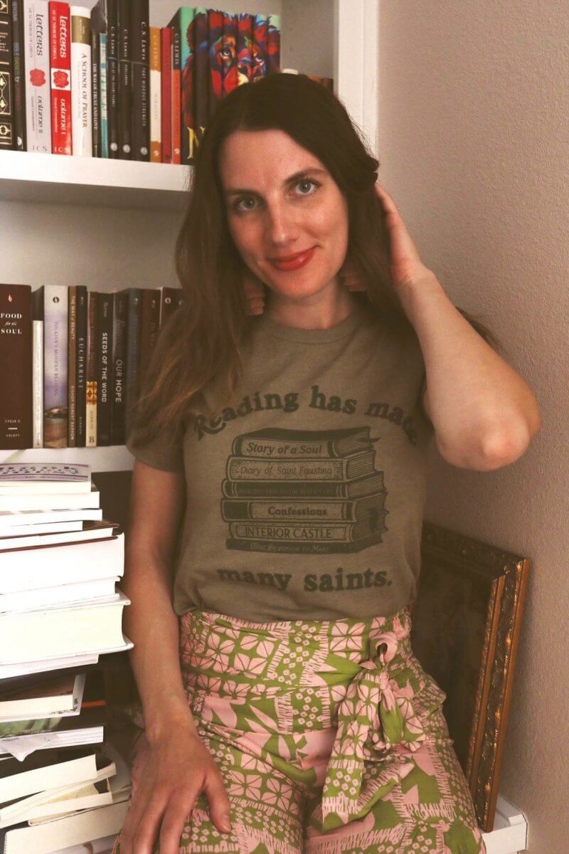 Woman wearing St. Josemaria Escriva tee with text 'Reading has made many saints' in a cozy library setting.