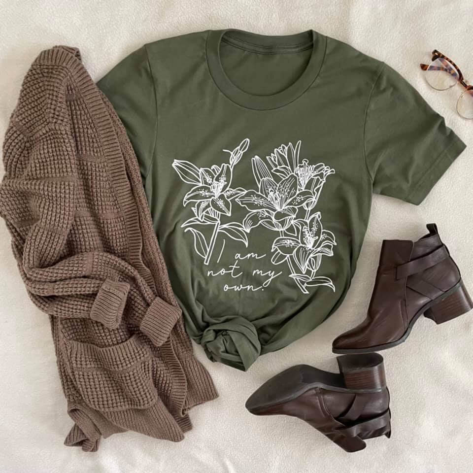 St. Kateri I Am Not My Own Tee in military green with floral design, paired with brown cardigan, ankle boots, and glasses on cozy surface.