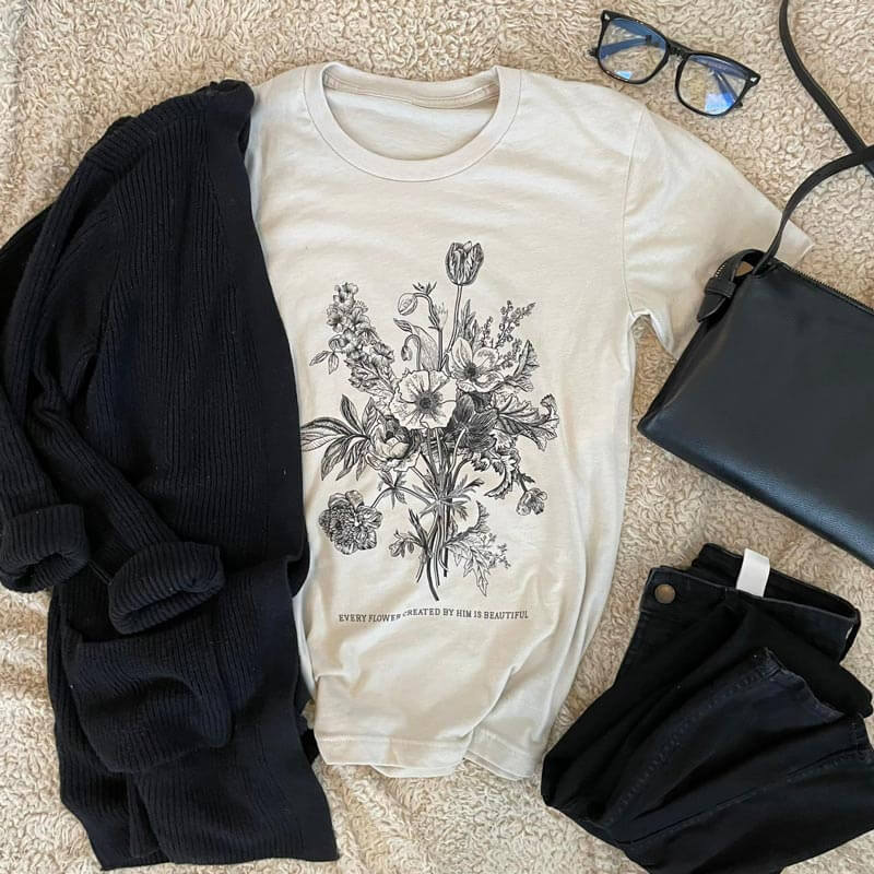 St. Therese de Lisieux Every Flower Tee with floral print, laid flat with black cardigan, bag, glasses, and pants.