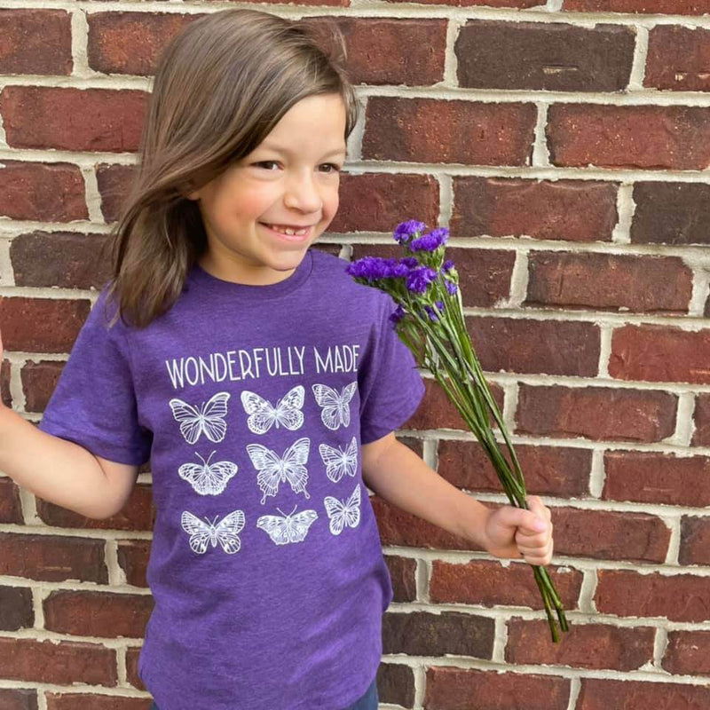 Toddler wearing purple 'Wonderfully Made' tee with butterfly design, holding purple flowers against brick wall.