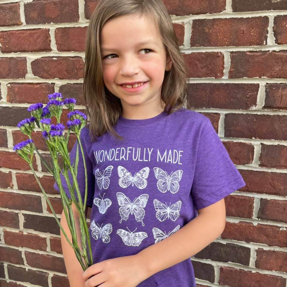 Toddler wearing 'Wonderfully Made' purple tee with butterfly design, holding purple flowers, standing against a brick wall.